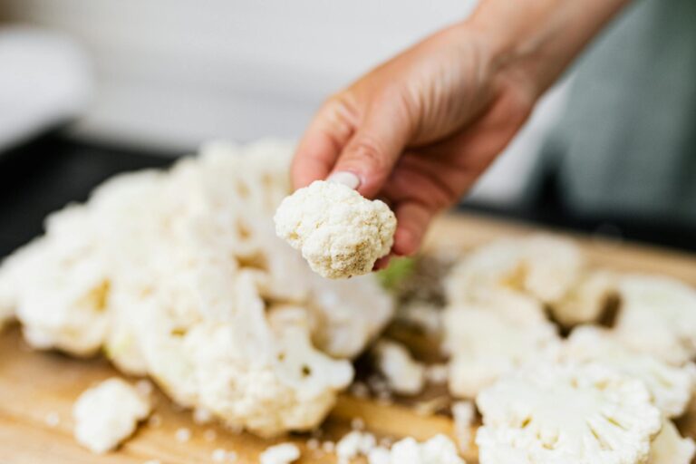 A hand holds a fresh cauliflower floret over a wooden board with scattered florets, showcasing a cooking preparation.
